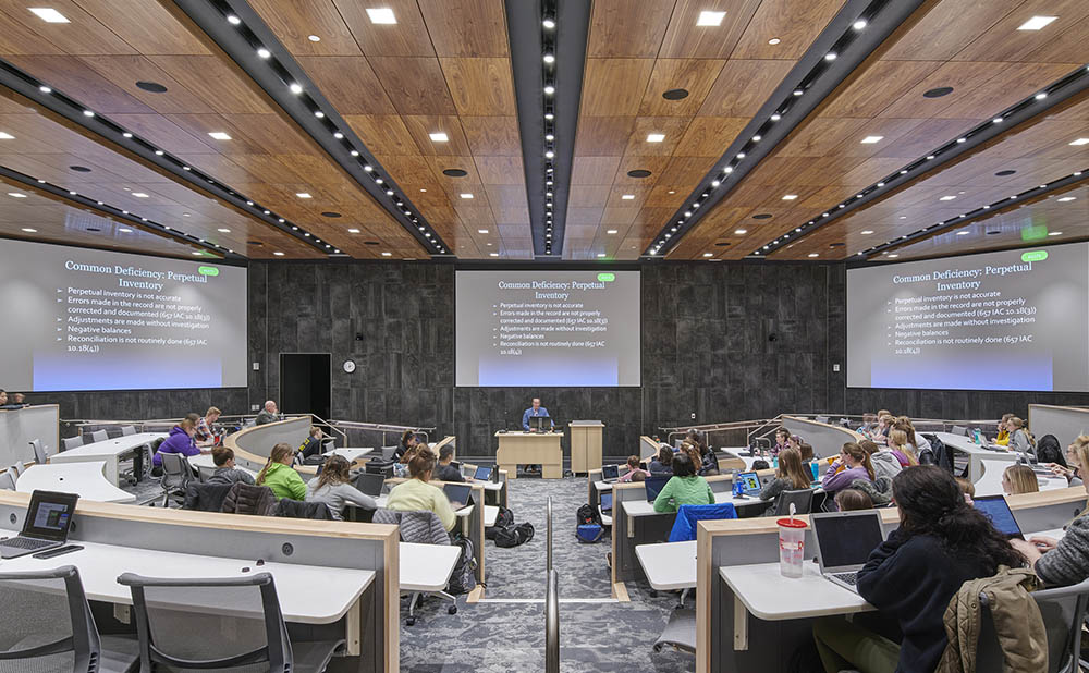 tiered classroom seating facing professor and three large screens